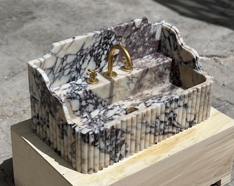 May include: A white and purple marble sink with a gold faucet and a fluted base. The sink is sitting on a wooden platform.