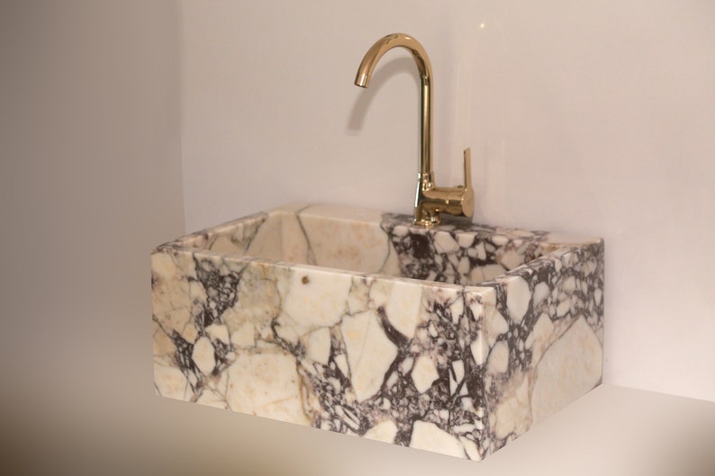 Pu&ograve; includere: Un lavabo in marmo bianco con un rubinetto dorato. Il lavabo &egrave; rettangolare ed &egrave; realizzato in pietra naturale con un motivo venato unico.