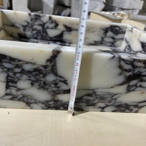 Peut inclure: Un &eacute;vier en marbre blanc avec des veines noires. L'&eacute;vier est rectangulaire et a un bord arrondi. Un ruban &agrave; mesurer est plac&eacute; le long du bord de l'&eacute;vier, montrant une mesure de 45,7 cm.