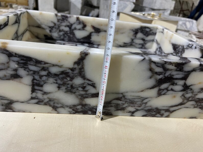 Peut inclure: Un &eacute;vier en marbre blanc avec des veines noires. L'&eacute;vier est rectangulaire et a un bord arrondi. Un ruban &agrave; mesurer est plac&eacute; le long du bord de l'&eacute;vier, montrant une mesure de 45,7 cm.