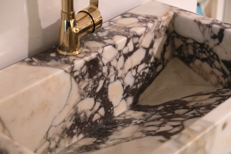 Pu&ograve; includere: Un lavandino in marmo bianco con venature marroni e nere. Il lavandino &egrave; rettangolare e ha un rubinetto dorato.