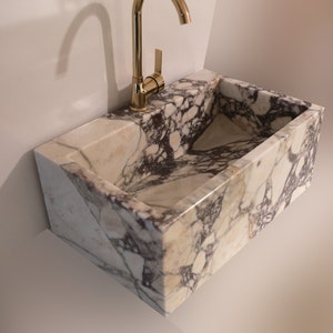 Pu&ograve; includere: Un lavabo in marmo bianco con un rubinetto dorato. Il lavabo &egrave; rettangolare e ha un bordo curvo. Il lavabo &egrave; montato a parete.