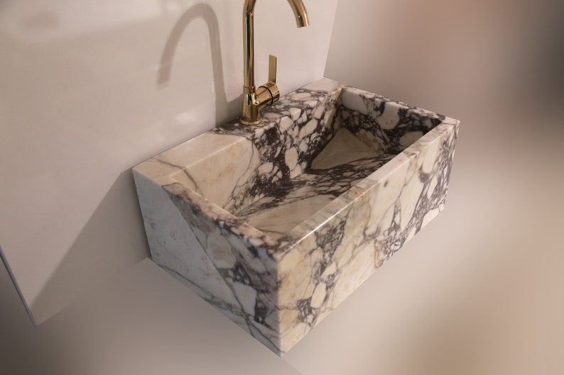 Pu&ograve; includere: Un lavabo in marmo bianco con un rubinetto dorato. Il lavabo &egrave; rettangolare e ha un bordo curvo. Il lavabo &egrave; montato a parete.