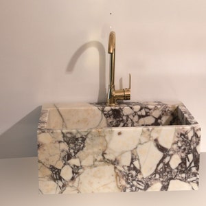 Pu&ograve; includere: Un lavabo in marmo bianco con un rubinetto dorato. Il lavabo &egrave; rettangolare ed &egrave; realizzato in pietra naturale. Il rubinetto &egrave; alto e ha un'unica maniglia.