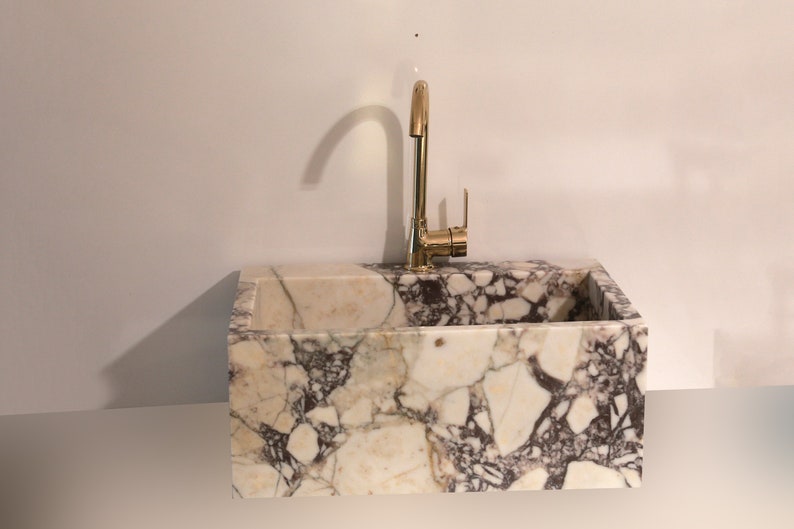 Pu&ograve; includere: Un lavabo in marmo bianco con un rubinetto dorato. Il lavabo &egrave; rettangolare ed &egrave; realizzato in pietra naturale. Il rubinetto &egrave; alto e ha un'unica maniglia.