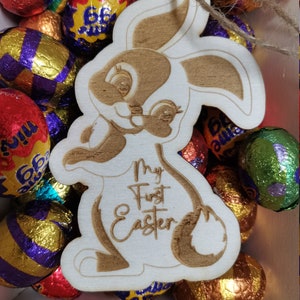 May include: A wooden bunny cutout with the text "My First Easter" engraved on it. The bunny is surrounded by colorful foil-wrapped chocolate Easter eggs.