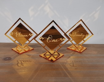 May include: Three gold mirrored table number holders with a diamond shape. The numbers "One", "Eleven", and "Twenty" are engraved on the holders.