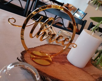 May include: Gold table number with the word "three" in a cursive font, on a wooden slice with a white candle and a glass vase with greenery.