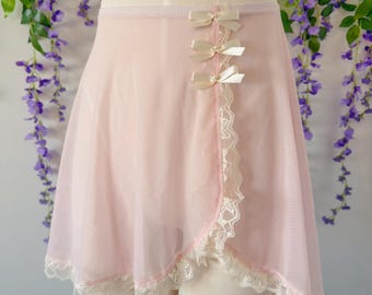 May include: A pale pink wrap skirt made of sheer mesh fabric featuring a scalloped white lace trim along the hem and front edge. The skirt is accented with three small cream-colored satin bows vertically aligned along the side closure.