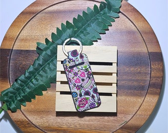 May include: A pink, white, and black key chain with a sugar skull design. The key chain is on a wooden surface with a green fern.