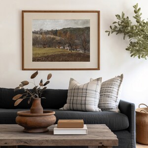 May include: A framed landscape painting with a brown wooden frame hangs above a dark gray sofa with plaid pillows. A wooden coffee table holds a wooden vase with dried leaves, a brown box, and a white box. A woven basket sits on the right.