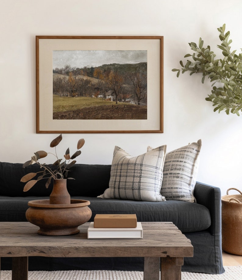 May include: A framed landscape painting with a brown wooden frame hangs above a dark gray sofa with plaid pillows. A wooden coffee table holds a wooden vase with dried leaves, a brown box, and a white box. A woven basket sits on the right.