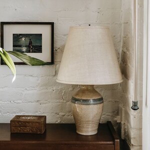 May include: A ceramic table lamp with a beige and blue patterned base and a white linen lampshade. The lamp is sitting on a dark brown wooden table.