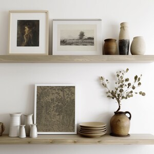 May include: Two wooden shelves with framed artwork, vases, and a ceramic pitcher with dried eucalyptus branches. The artwork includes a black and white landscape print, a black and white drawing of trees, and a portrait of a woman. The vases are in various shapes and sizes, including a tall, narrow vase, a round vase, and a squat vase. The shelves are against a white wall.