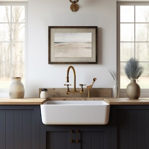 May include: A farmhouse kitchen sink with a white porcelain basin and a brass faucet. The sink is set into a dark blue cabinet with a wooden countertop. A framed seascape painting hangs on the wall above the sink.