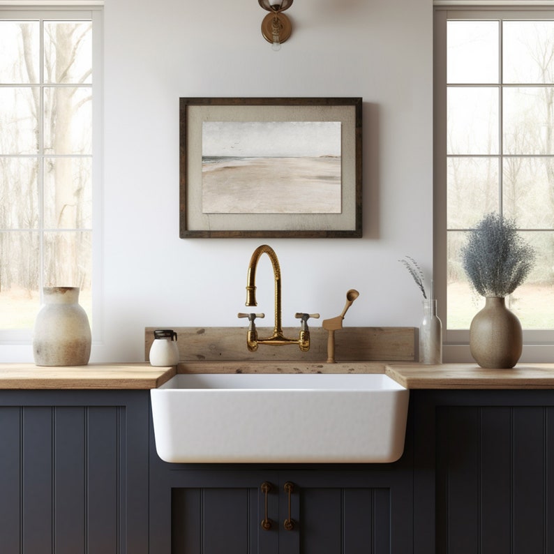 May include: A farmhouse kitchen sink with a white porcelain basin and a brass faucet. The sink is set into a dark blue cabinet with a wooden countertop. A framed seascape painting hangs on the wall above the sink.