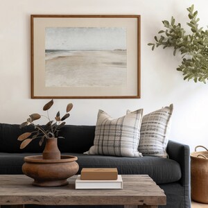 May include: A dark gray sofa with two pillows, one with a black and white plaid pattern and the other with a gray and white striped pattern. A wooden coffee table with a brown box and a white book on top. A vase with dried branches sits on the table. A framed watercolor painting of a beach scene hangs on the wall behind the sofa.