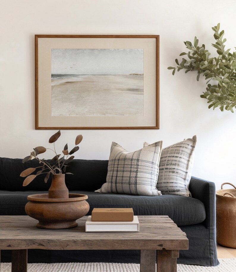 May include: A dark gray sofa with two pillows, one with a black and white plaid pattern and the other with a gray and white striped pattern. A wooden coffee table with a brown box and a white book on top. A vase with dried branches sits on the table. A framed watercolor painting of a beach scene hangs on the wall behind the sofa.