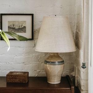 May include: A ceramic lamp with a beige and blue glaze, featuring a white linen lampshade. The lamp is sitting on a wooden table next to a small wooden box.