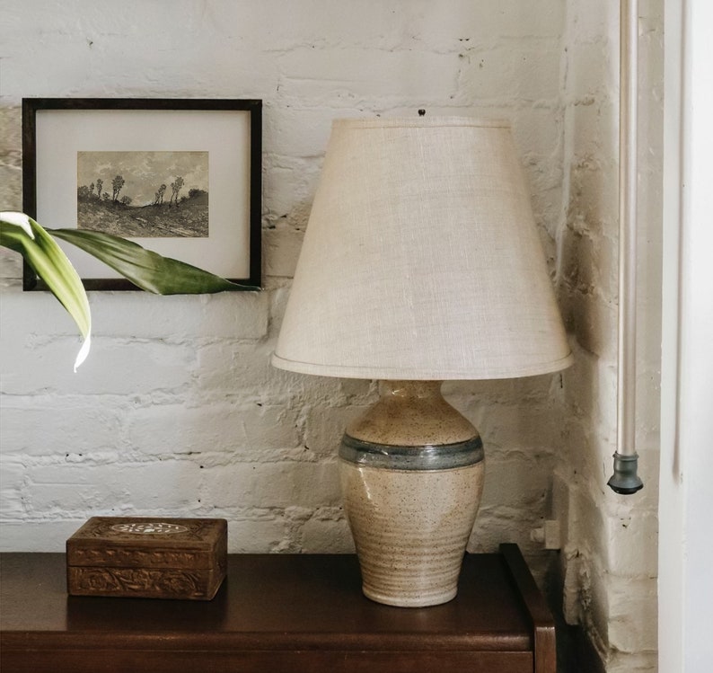 May include: A ceramic lamp with a beige and blue glaze, featuring a white linen lampshade. The lamp is sitting on a wooden table next to a small wooden box.