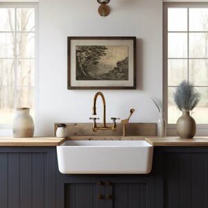 May include: A white farmhouse kitchen sink with a brass faucet and a wooden countertop. The sink is in front of a window with a view of trees and a wooden framed print of a landscape.