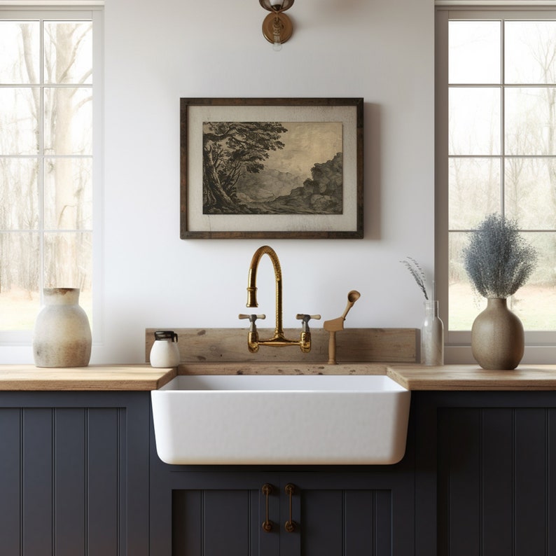May include: A white farmhouse kitchen sink with a brass faucet and a wooden countertop. The sink is in front of a window with a view of trees and a wooden framed print of a landscape.