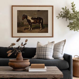 以下が含まれることがあります： 茶色と白の馬が野原に立っている絵画が額装されています。絵画は、白の壁に、2つのクッションのある濃い灰色のソファの上にかけられています。ソファの前に、木製のコーヒーテーブルがあり、木製のボウルと本が置かれています。