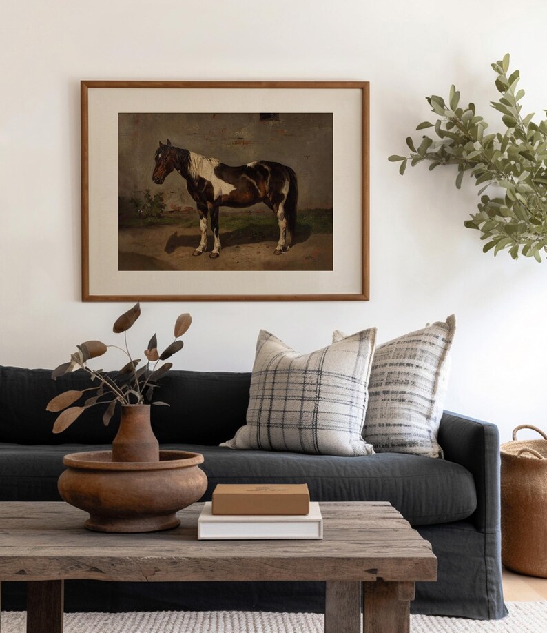 以下が含まれることがあります： 茶色と白の馬が野原に立っている絵画が額装されています。絵画は、白の壁に、2つのクッションのある濃い灰色のソファの上にかけられています。ソファの前に、木製のコーヒーテーブルがあり、木製のボウルと本が置かれています。