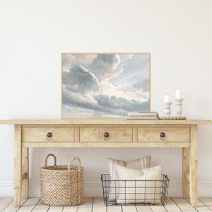 May include: A wooden console table with three drawers and a framed painting of a cloudy sky above a field. The table has a natural wood finish and the painting is in a light brown frame.