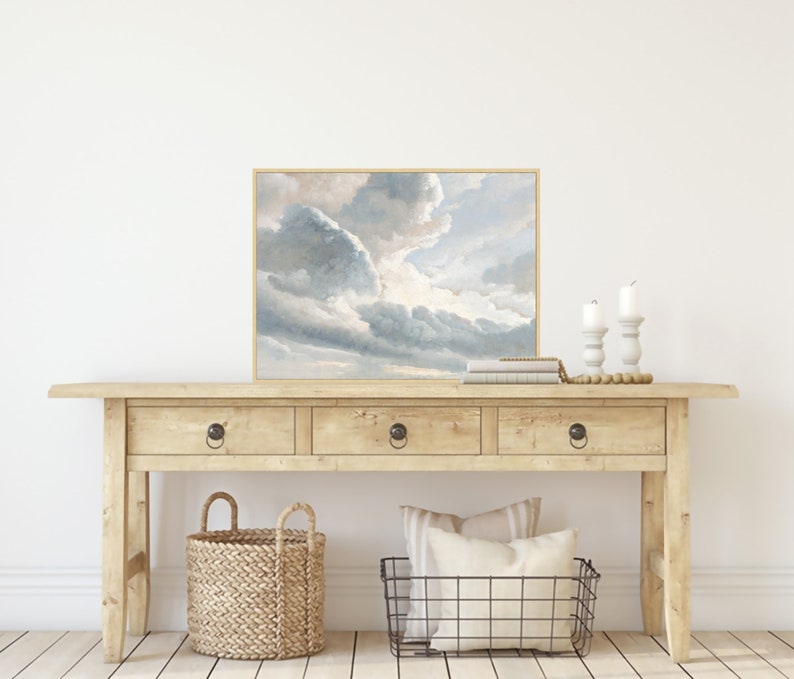 May include: A wooden console table with three drawers and a framed painting of a cloudy sky above a field. The table has a natural wood finish and the painting is in a light brown frame.