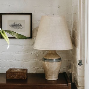 Op de afbeelding: Een keramische lamp met een beige en blauwe glazuur, met een witte linnen lampenkap. De lamp staat op een donkerbruin houten oppervlak.