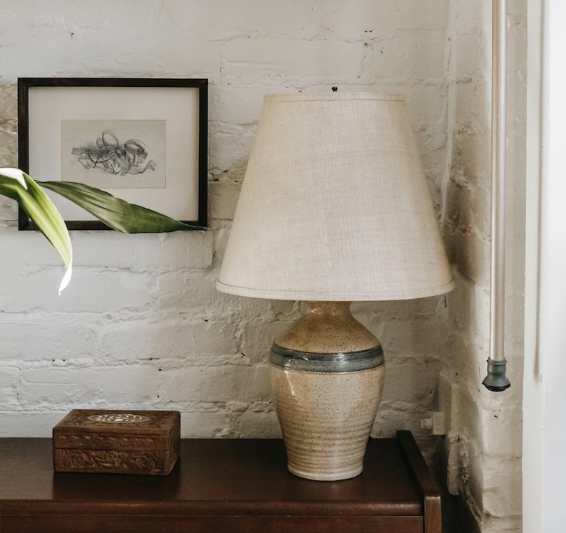 Op de afbeelding: Een keramische lamp met een beige en blauwe glazuur, met een witte linnen lampenkap. De lamp staat op een donkerbruin houten oppervlak.