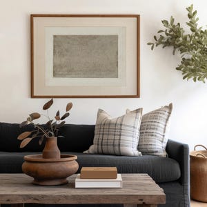 May include: A dark gray linen sofa with two throw pillows, one with a black and white plaid pattern and the other with a gray and white striped pattern. A wooden coffee table with a brown wooden bowl and a white book with a brown box on top sits in front of the sofa. A potted plant with green leaves is in the background.