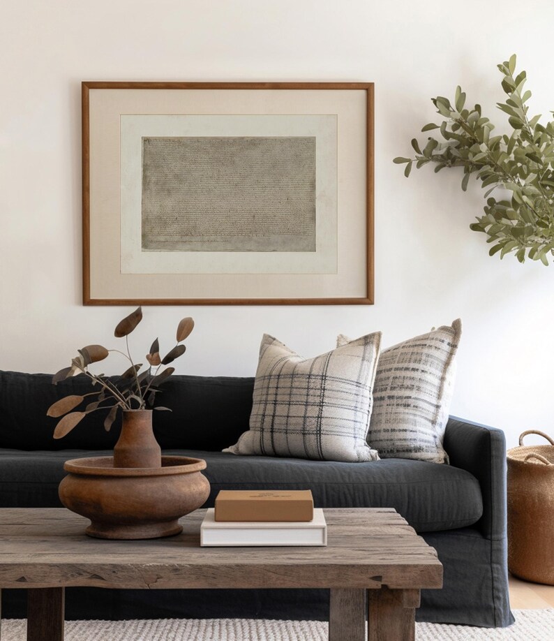 May include: A dark gray linen sofa with two throw pillows, one with a black and white plaid pattern and the other with a gray and white striped pattern. A wooden coffee table with a brown wooden bowl and a white book with a brown box on top sits in front of the sofa. A potted plant with green leaves is in the background.