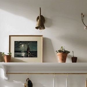 May include: A white shelf with a brass rail and hooks hangs on a white wall. A framed print of a painting depicting a person walking a dog on a beach at night is on the shelf. A brass wall sconce with a bell-shaped shade is mounted on the wall above the shelf. A potted plant sits on the shelf to the right of the print. A vase with a branch of white flowers sits on the shelf to the right of the plant.