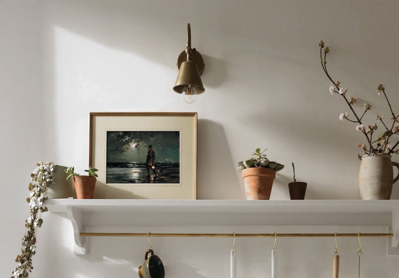 May include: A white shelf with a brass rail and hooks hangs on a white wall. A framed print of a painting depicting a person walking a dog on a beach at night is on the shelf. A brass wall sconce with a bell-shaped shade is mounted on the wall above the shelf. A potted plant sits on the shelf to the right of the print. A vase with a branch of white flowers sits on the shelf to the right of the plant.