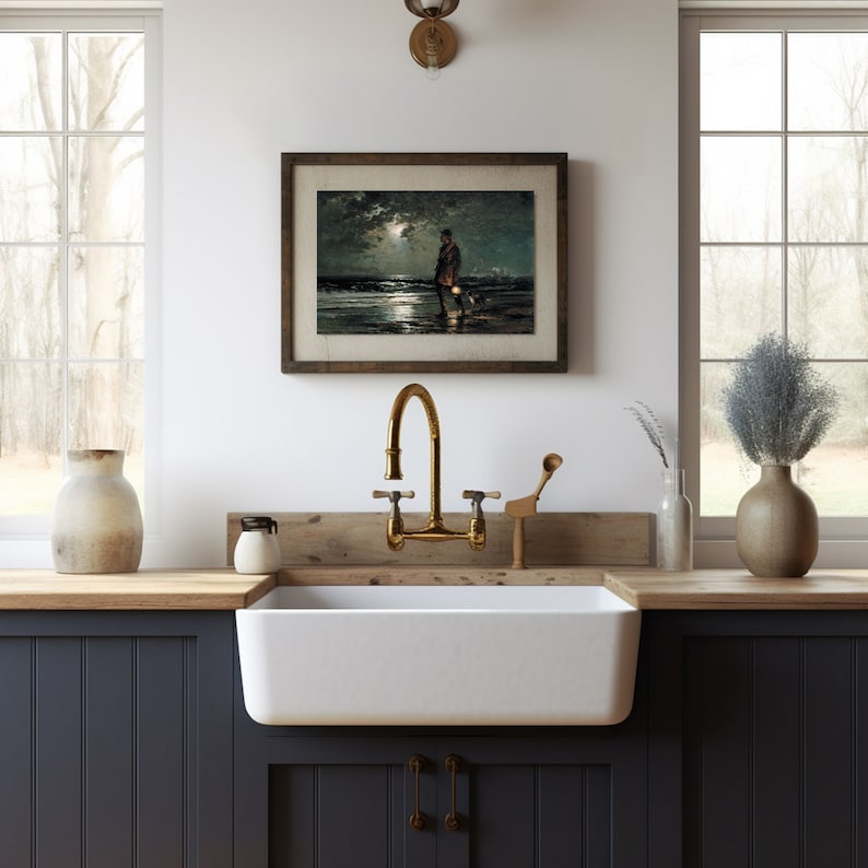 May include: A white farmhouse kitchen sink with a gold faucet and a framed black and white print of a person walking a dog on a beach at night.