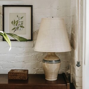 May include: A beige ceramic table lamp with a cream-colored lampshade. The lamp has a speckled finish with a blue stripe. A framed botanical print and a wooden box are also visible.