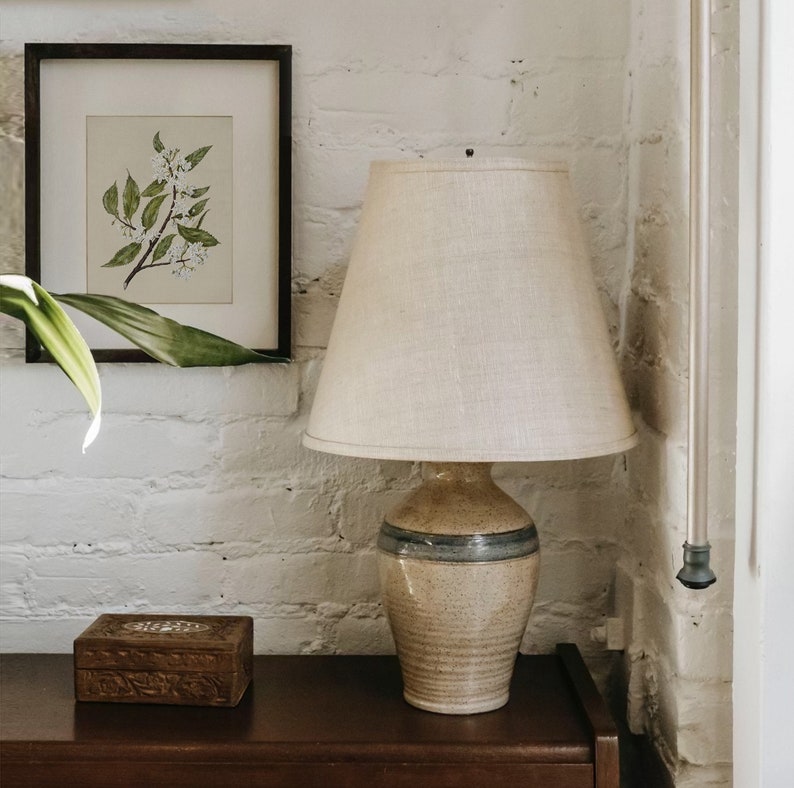 May include: A beige ceramic table lamp with a cream-colored lampshade. The lamp has a speckled finish with a blue stripe. A framed botanical print and a wooden box are also visible.
