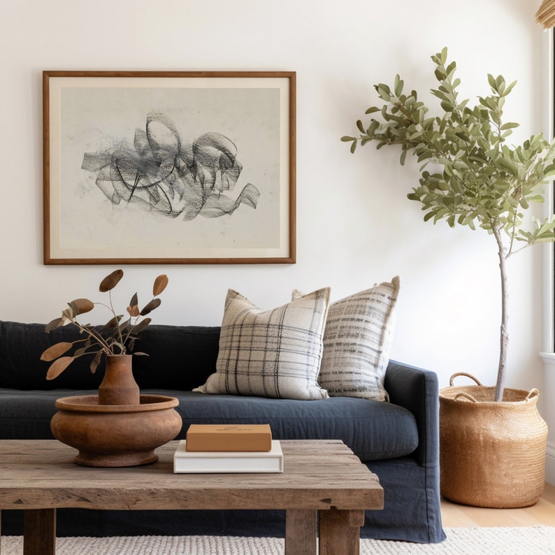Op de afbeelding: Een donkerblauwe bank met twee kussens, een houten salontafel, een vaas met gedroogde takken en een geweven mand met een plant in een pot. De muur achter de bank heeft een ingelijst abstract zwart-wit ontwerp.