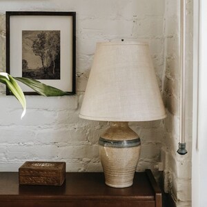 May include: A ceramic table lamp with a beige and blue patterned base and a white linen lampshade. The lamp is sitting on a dark brown wooden table.