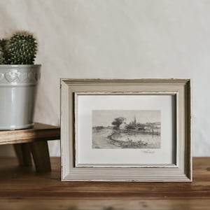 May include: A framed black and white sketch of a landscape scene. The artwork is in a distressed, light gray wooden frame. A small cactus in a gray pot sits to the left, and a ceramic mug is on the right.