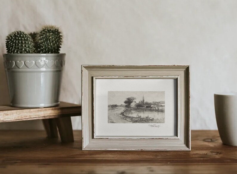 May include: A framed black and white sketch of a landscape scene. The artwork is in a distressed, light gray wooden frame. A small cactus in a gray pot sits to the left, and a ceramic mug is on the right.