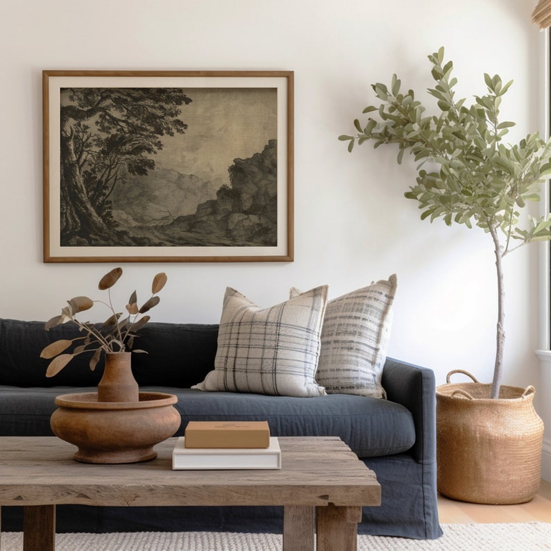 May include: A dark blue sofa with two pillows, one with a plaid pattern and one with a striped pattern. A wooden coffee table with a vase of dried branches, a book, and a box on top. A large potted plant with green leaves in a woven basket is in the corner of the room. A framed black and white landscape print hangs on the wall.