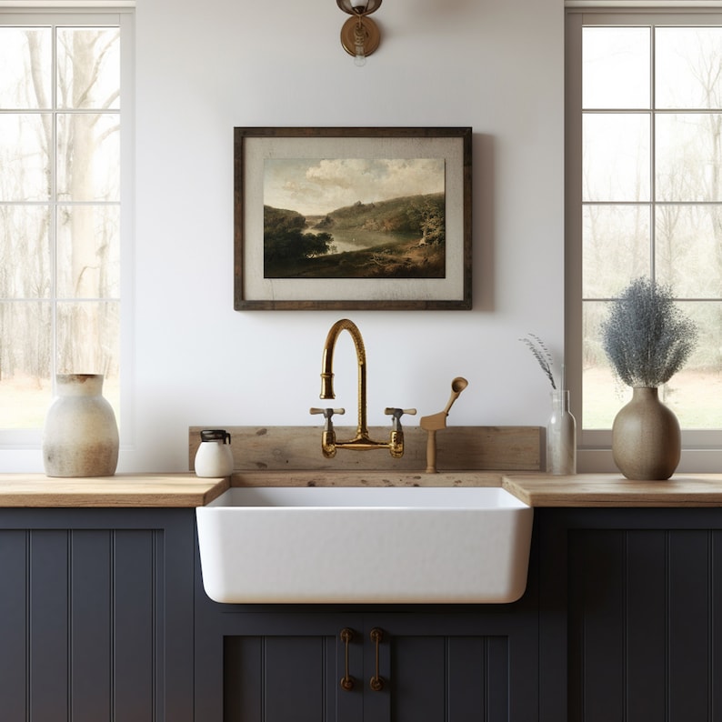 Pu&ograve; includere: Un lavello da cucina bianco in stile farmhouse con un rubinetto in ottone e un piano di lavoro in legno. Il lavello &egrave; posizionato contro armadietti blu scuro. Un dipinto paesaggistico incorniciato &egrave; appeso al muro sopra il lavello.