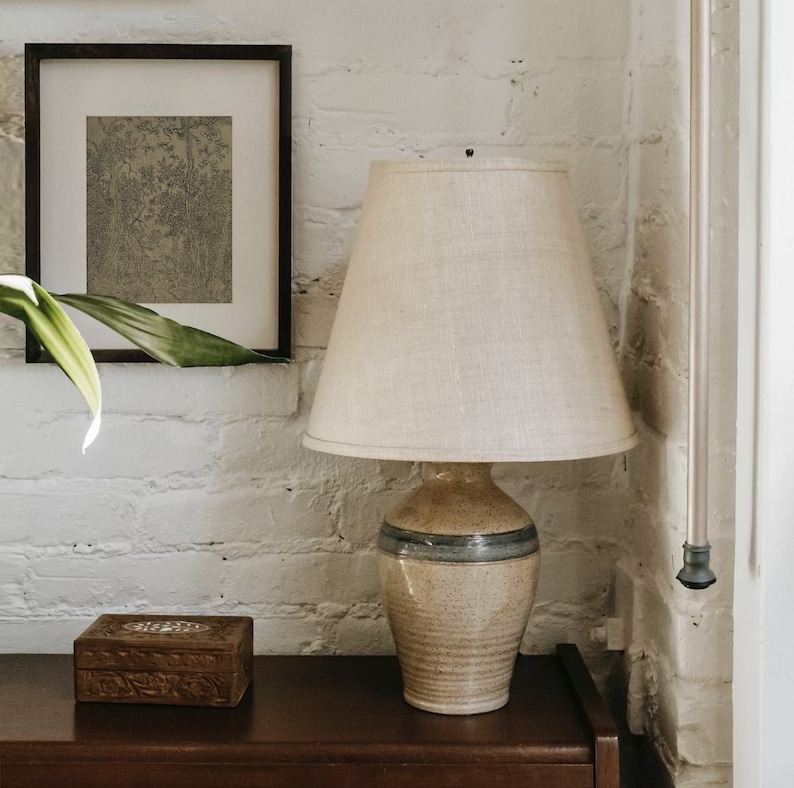May include: A ceramic table lamp with a beige linen lampshade. The lamp base is a speckled beige with a blue band around the middle. The lamp is sitting on a dark wood table next to a wooden box.