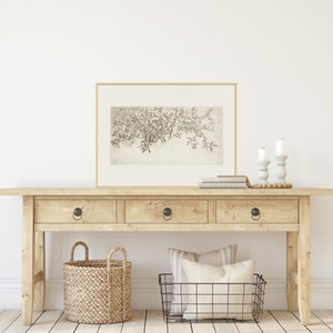 Op de afbeelding: Een lichte houten consoletafel met drie laden en zwarte metalen knoppen. De tafel is versierd met een ingelijst zwart-wit botanisch drukwerk, twee witte kandelaars, een stapel boeken, een geweven mand en een draadmand.