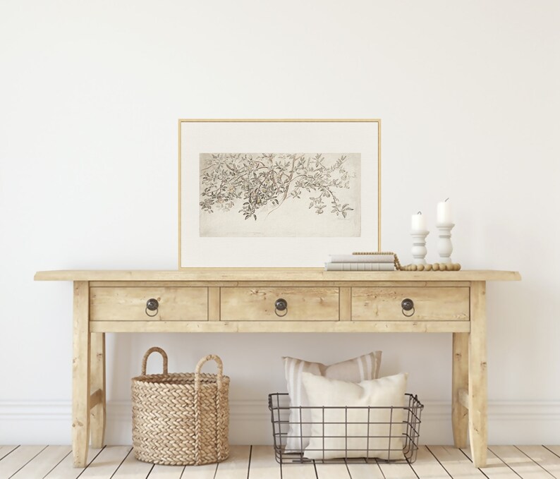 Op de afbeelding: Een lichte houten consoletafel met drie laden en zwarte metalen knoppen. De tafel is versierd met een ingelijst zwart-wit botanisch drukwerk, twee witte kandelaars, een stapel boeken, een geweven mand en een draadmand.