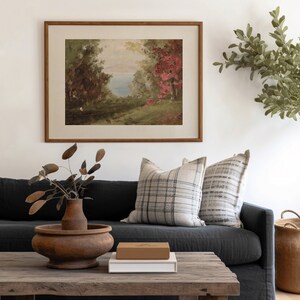 May include: A framed landscape painting depicting a lake scene with trees and a small cottage in the distance. The painting is in a wooden frame and is hanging on a white wall.