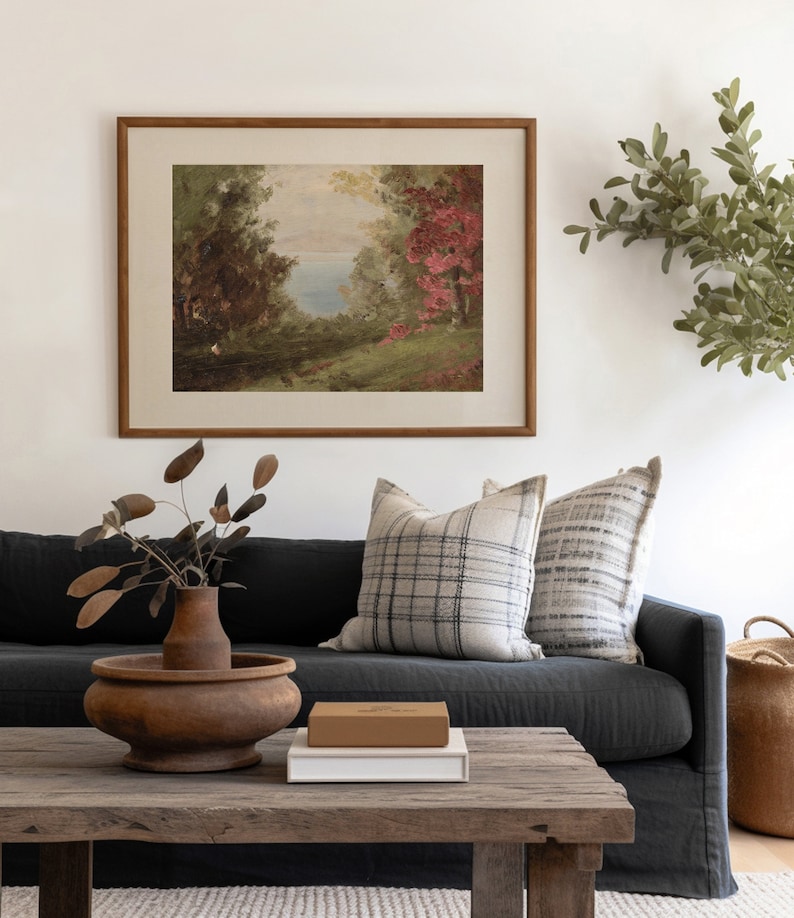 May include: A framed landscape painting depicting a lake scene with trees and a small cottage in the distance. The painting is in a wooden frame and is hanging on a white wall.